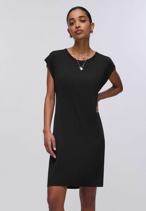 Donna in piedi con una mano sul fianco, indossa un semplice vestito nero senza maniche, orecchini a cerchio dorati e collane a strati.