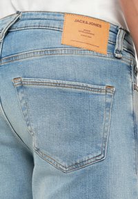 Gai zila džinsu bikses ar ādas uzlīmi, kurā rakstīts "JACK & JONES", uz jostas, ar aizmugures kabatām, kurām ir kontrastējoša dūriena.
