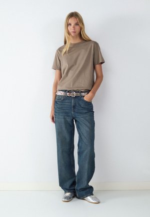 Frau mit langen blonden Haaren steht vor einer weißen Wand und trägt ein taupefarbenes T-Shirt, lockere blaue Jeans, einen silbernen Gürtel und silberne spitze Schuhe.