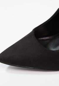 Escarpin en daim noir à bout pointu avec empeigne basse et semelle intérieure lisse sur fond blanc.