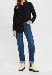 Camicia nera a maniche lunghe con colletto, abbinata a jeans blu e stivaletti beige alla caviglia con pannelli elastici neri e suole testurizzate.