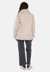 Giacca in pelliccia ecologica beige con ampio colletto, vestibilità strutturata, tasche laterali e orlo dritto, indossata sopra jeans scuri e sneakers bianche.