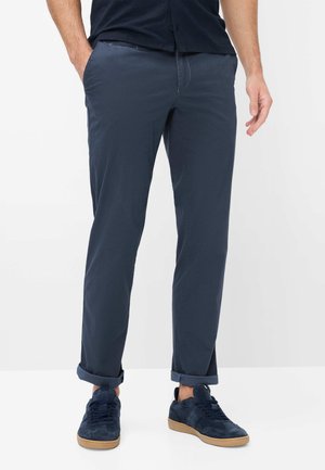 Mann mit hochgekrempelten dunkelblauen Hosen und passenden Sneakers, stehend mit einer Hand in der Tasche vor einfachem Hintergrund.