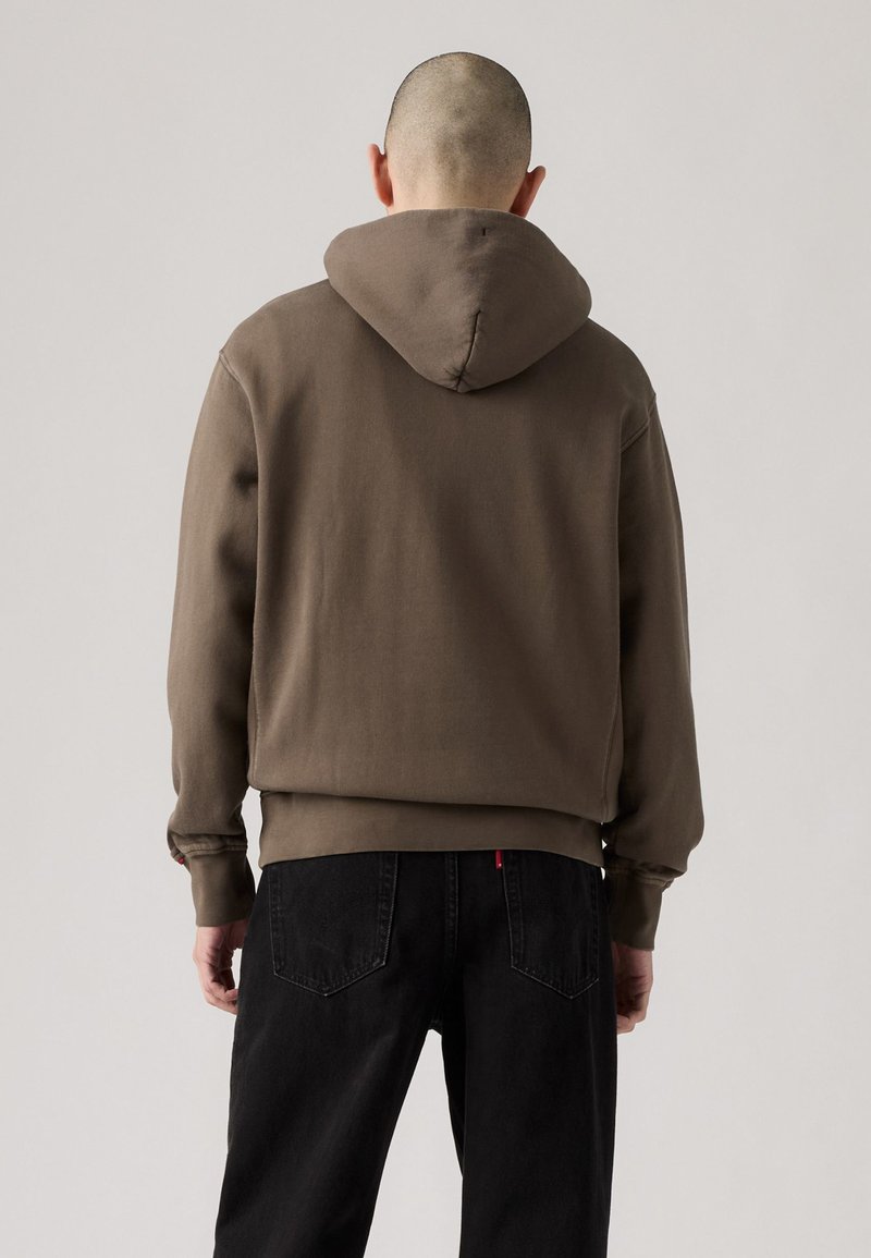 Brauner Hoodie mit Kordelzugkapuze, gerippten Bündchen und Saum. Getragen mit schwarzen Jeans, die einen lässigen Schnitt und eine weiche Textur zeigen.