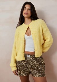 Giovane donna che indossa un cardigan giallo peloso su una canotta bianca e pantaloni corti con stampa leopardata, in piedi contro un muro beige.