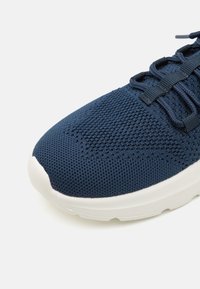 Marineblauwe sneaker met een gestructureerd mesh bovenwerk, witte rubberen zool en platte veters. Kenmerkend is de ronde neus en de verstevigde stikseldetails.