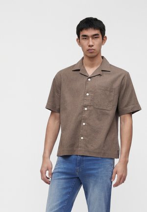 Jeune homme debout, portant une chemise marron à manches courtes avec une poche poitrine et un jean bleu clair sur fond uni.