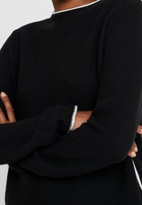 Svart tröja med rund hals, långa ärmar och vit accent längs halsringningen och ärmsluten. Texturerad tyg med en figursydd design.