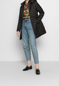 Manteau matelassé noir avec capuche, associé à une chemise florale colorée et un jean taille haute bleu clair. Des mocassins noirs complètent la tenue.