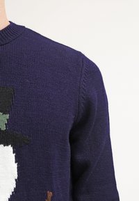 Suéter de punto morado con cuello redondo que presenta un diseño de muñeco de nieve en blanco, negro, verde y marrón. Tejido texturizado con un ligero estiramiento.