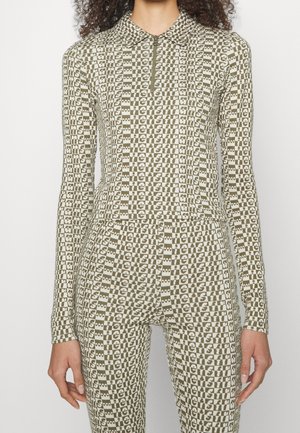 T-shirt ajusté à manches longues en vert olive avec un col zippé. Le tissu présente un motif de damier répétitif avec des blocs de texte alternés.