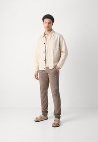 Junger Mann in beiger Jacke, hellem Pullover, braunen Hosen und beigen Sandalen, steht mit einer Hand in der Tasche vor einem weißen Hintergrund.