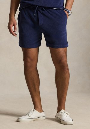 Tmavomodré mäkké teplákové šortky s elastickým pásom a šnúrkou na stiahnutie, s bočnými vreckami a malým logom na leme. Nosené s bielymi teniskami.