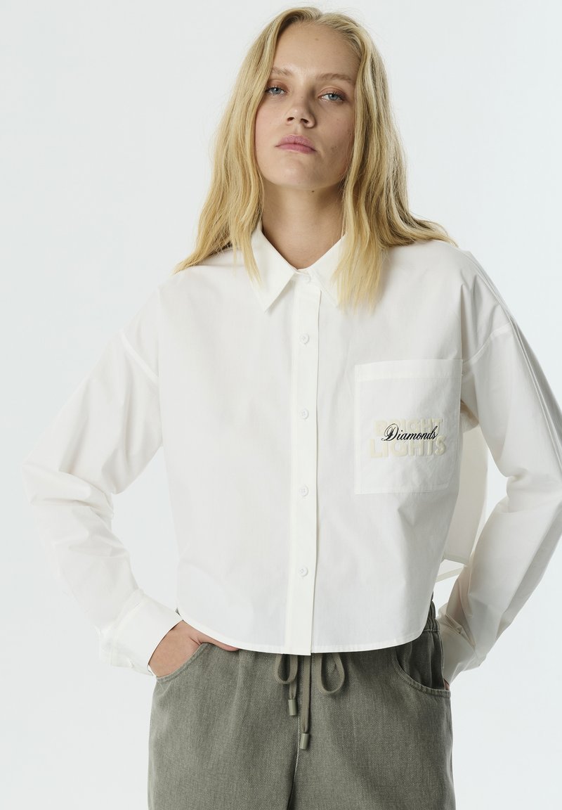 White cropped button-up shirt made of cotton, featuring a chest pocket with embroidered "Diamonds" text. Long sleeves with buttoned cuffs.