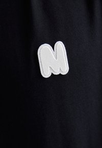 Sort stof med en hvid, tekstureret, broderet bogstav "M" patch, der har en let forhøjet kant og en glat inderside.