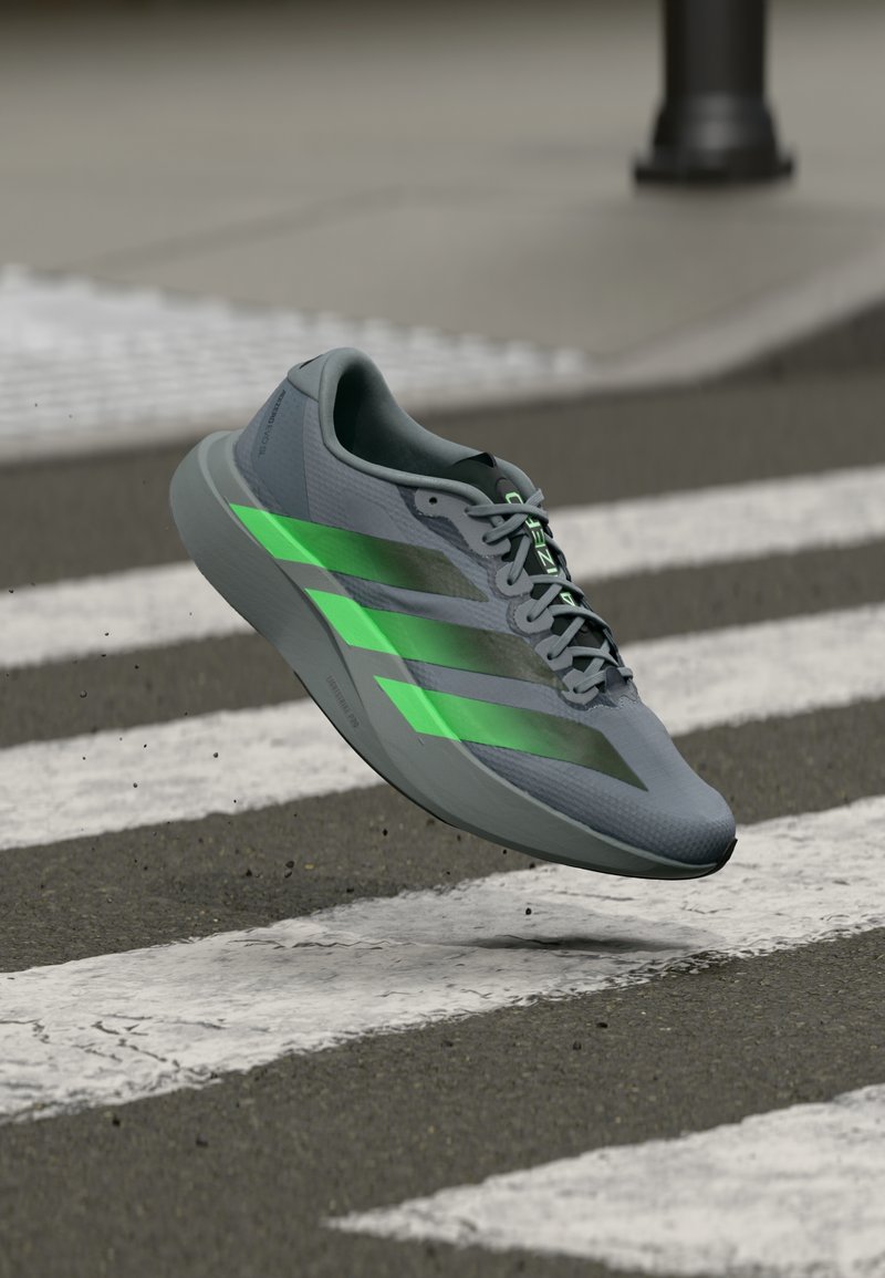 Chaussure de sport grise avec des bandes accentuées vert néon, une tige texturée et une semelle rembourrée, présentée en l'air au-dessus d'un passage piéton.
