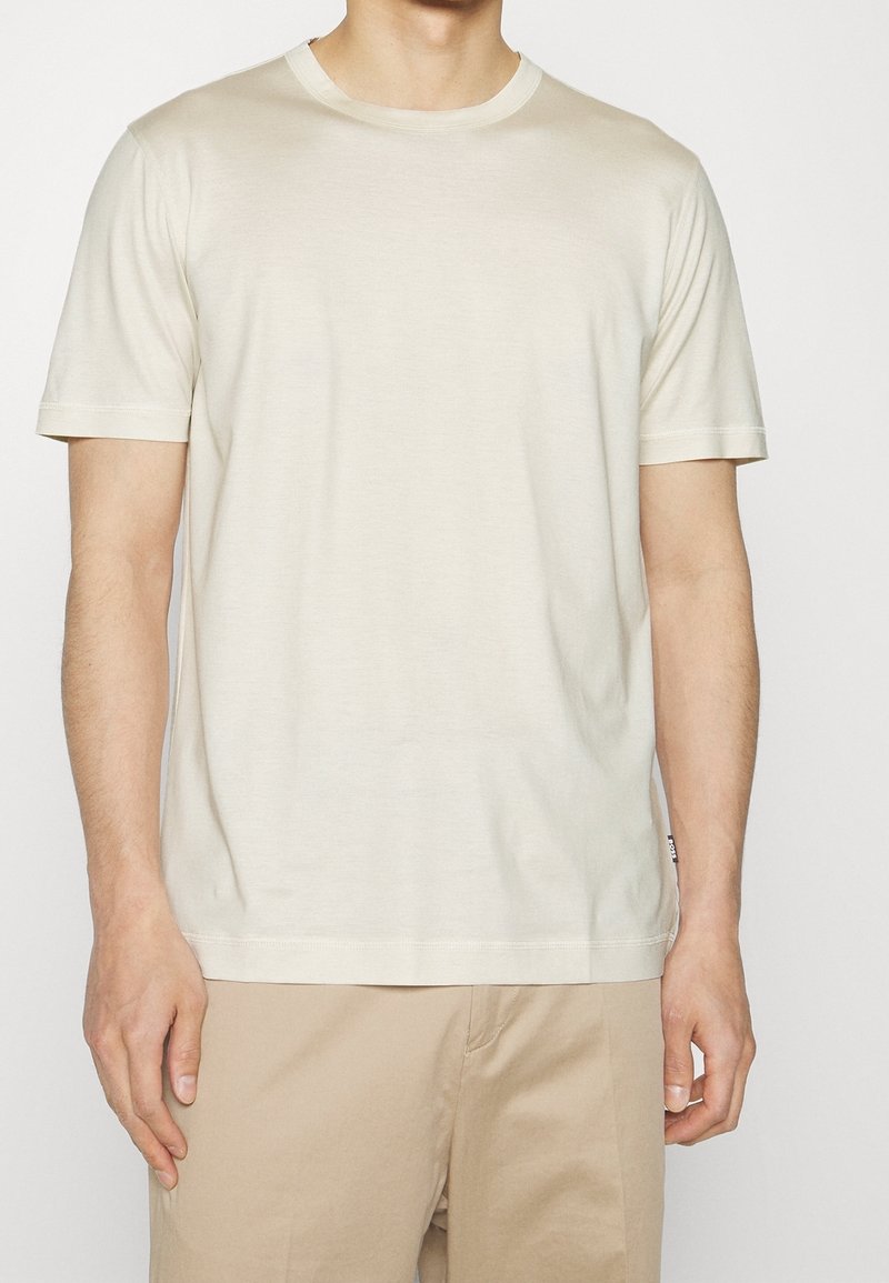 Homme portant un t-shirt uni à manches courtes de couleur blanc cassé avec un col rond et un pantalon beige, debout devant un fond uni.