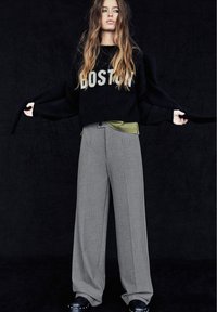 Černý svetr s nápisem "BOSTON" v krémové barvě, oblečený přes zelenou košili. Šedé široké kalhoty s hladkou texturou a knoflíkovým detailem. Černé dřeváky.