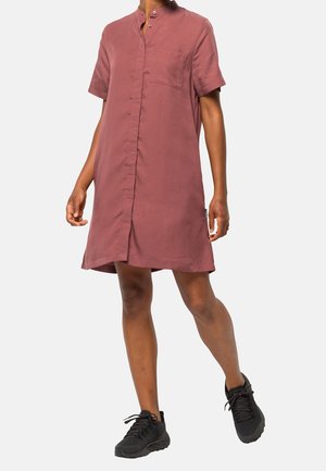 Kvinde iført en kortærmet, knælængde kjole i støvet rosa med sorte sneakers, gående mod en ensfarvet lys baggrund.