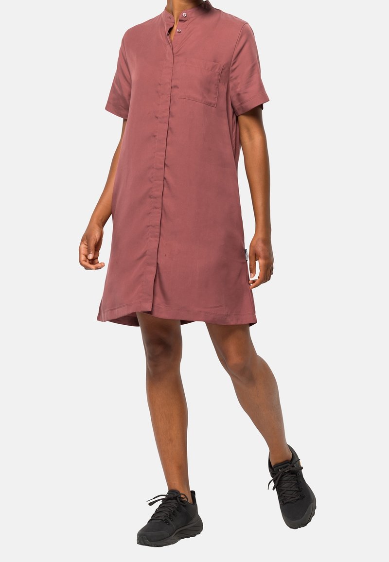 Mujer con un vestido camisa de manga corta hasta la rodilla en color rosa polvo y zapatillas negras, caminando sobre un fondo claro y liso.