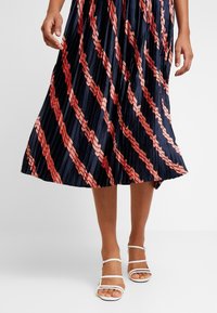 Person bär en plisserad marinblå kjol med diagonala röda och rosa ränder, tillsammans med vita sandaler med öppet tåparti och remmar.