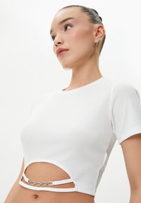 Hvid ribbet kortærmet crop top med udskæring og dekorative sølvkæde-detaljer i taljen. Glat tekstur.