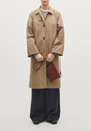 Cappotto oversize color cammello con maniche ampie, chiusura con un unico bottone, indossato sopra un maglione grigio e pantaloni scuri, mentre tiene una piccola borsa marrone.