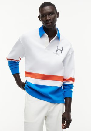 Mand iført hvid, blå og orange langærmet polo med "Tommy Hilfiger Marina Club"-logo, kombineret med hvide bukser, poserer mod en hvid baggrund.
