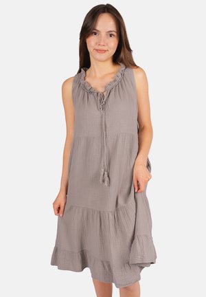 Femme portant une robe sans manches grise claire à volants avec col froncé et lien à pompons, debout devant un fond blanc uni.