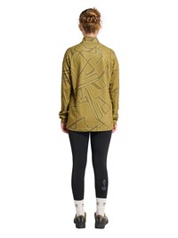 Kvinde stående med ryggen til, iført en olivengrøn mønstret bluse med lange ærmer, sorte leggings med teksten "ATW", hvide sokker og sorte sko.