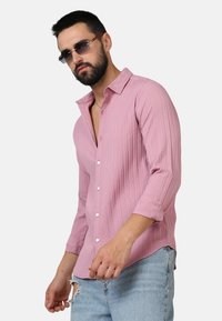 Camicia a maniche lunghe rosa chiaro con texture a righe verticali, chiusura con bottoni e maniche arrotolate, abbinata a jeans in denim strappati.