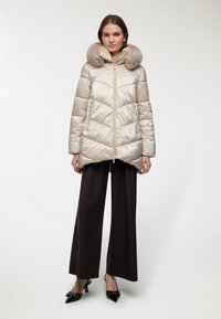 Giacca imbottita beige con cappuccio in pelliccia sintetica, dotata di zip frontale, trapuntatura a chevron e tasche laterali, abbinata a pantaloni larghi marroni.