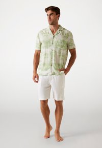 Kurzarmhemd in Hellgrün mit einem Palmenmuster, aus leichtem Stoff gefertigt, kombiniert mit weißen Leinen-Shorts.