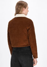 Giacca di velluto a coste marrone con colletto in shearling crema, design cropped e polsini a coste. La vista posteriore mostra dettagli e cuciture. Jeans neri.