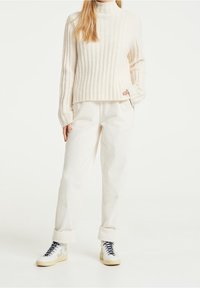 Pull en maille côtelée crème avec col haut, associé à un pantalon en velours côtelé écru. Des baskets blanches à lacets bleus complètent le look.