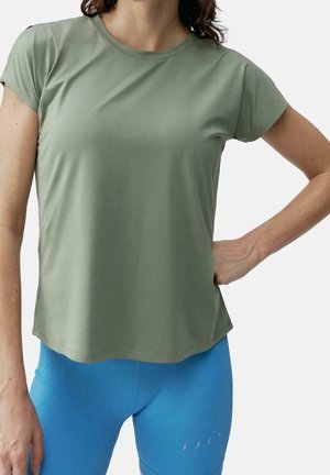 Vrouw draagt een lichtgroen T-shirt met korte mouwen en felblauwe leggings, staand met één hand op haar heup tegen een effen achtergrond.