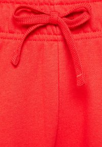 Červené tepláky s texturovanou tkaninou, elastickým pasem a tkaničkou. Detailní záběry zvýrazňují švy a uzlové detaily.