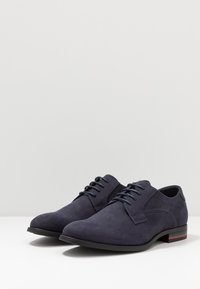 Chaussures en suède bleu marine à lacets avec une finition lisse, un bout rond, des détails de couture subtils et un accent rouge contrastant au talon.