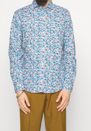 Man draagt een overhemd met knopen met een blauw, roze en zwart bladerenpatroon, gecombineerd met mosterdkleurige broek en een zilveren armband om de linkerpols.