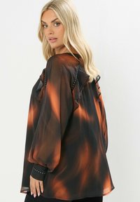 Blusa transparente de manga larga en negro con patrones naranjas, con volantes en la parte delantera y puños con tachuelas. Tejido ligero y fluido.