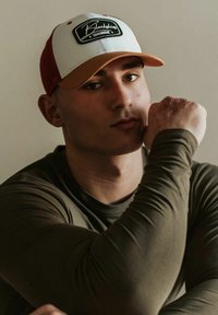 Béžová čiapka s bielym predným panelom a oranžovým šiltom, s čiernou nášivkou. Olivovo zelená dlhá rukávová košeľa, hladká textúra, detailný záber osoby.