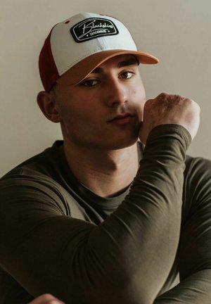 Baseball cepure ar baltu priekšpusi un oranžu malā, kurai pievienots melns ielāps. Olīvu zaļa garā piedurkne krekls, gluda tekstūra, tuvplāns uz cilvēku.