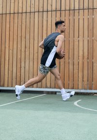 Sportiška sportinė apranga, apimanti juodai baltą be rankovių marškinėlį, raštuotas šortus ir baltus sportinius batelius, fone su mediniu paviršiumi.