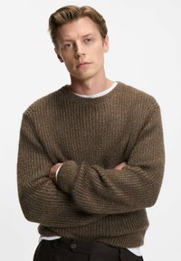 Maglione marrone a maglia testurizzata con collo a giro e vestibilità ampia, con polsini e orlo a coste. Indossato sopra una camicia bianca.