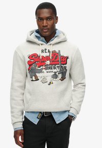 Felpa grigia con stampa grafica frontale, che presenta personaggi in stile cartone animato. Indossata sopra una camicia di jeans blu chiaro, con pantaloni neri.