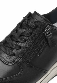 Sneaker nera in pelle con finitura testurizzata, caratterizzata da un dettaglio con zip e lacci neri piatti. La suola bianca accentua il design.