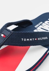 Infradito blu navy con cinturino intrecciato, caratterizzati dal testo bianco "TOMMY HILFIGER". La soletta presenta accenti di design in rosso, bianco e blu.