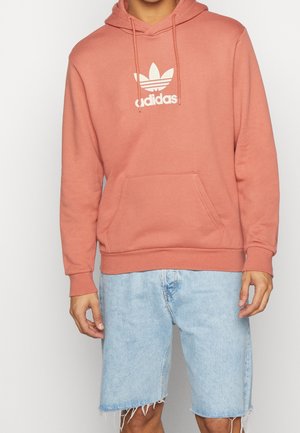 Sudadera con capucha en rosa empolvado con el logo de trébol blanco de Adidas, material de algodón, bolsillo frontal y puños de canalé. Llevada con pantalones cortos de mezclilla azul claros.