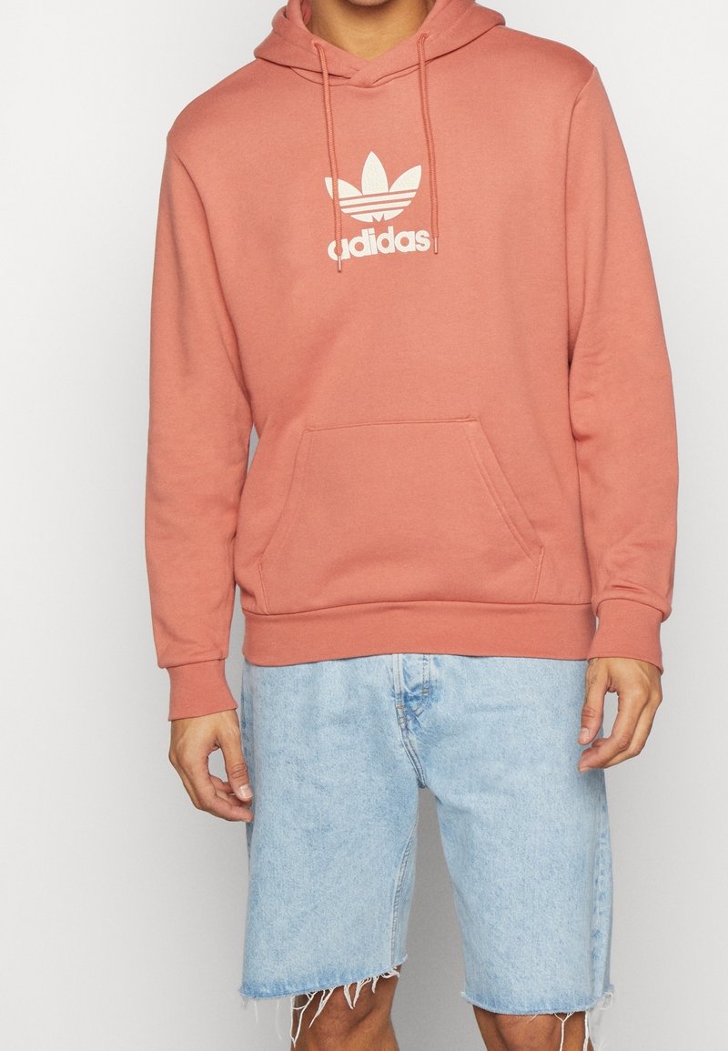 Hoodie i dammros med vit Adidas trefoil-logotyp, tillverkat av bomull, med framfickor och ribbade ärmslut. Bärs tillsammans med ljusblå denimshorts.