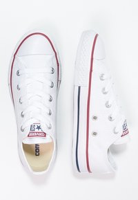 Vita canvas sneakers med gummisula, röd randning och en strukturerad tåhuvud. Har silverfärgade öglor och en varumärkesetikett på plösen.